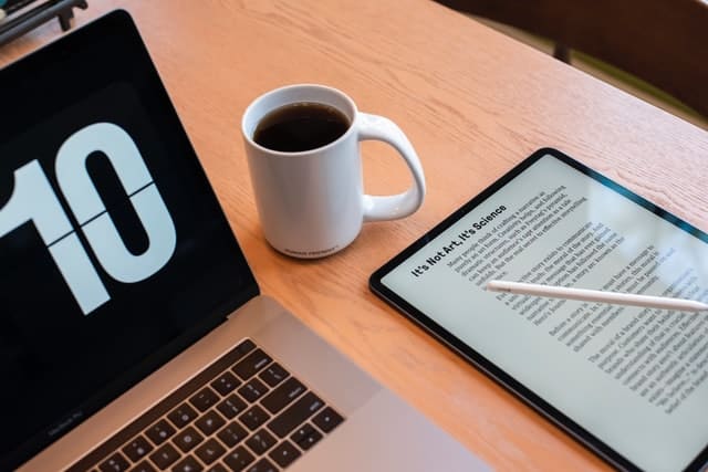 Table showing corner of a laptop, a coffee cup, and an ipad tablet with an opened ebook on the screen.