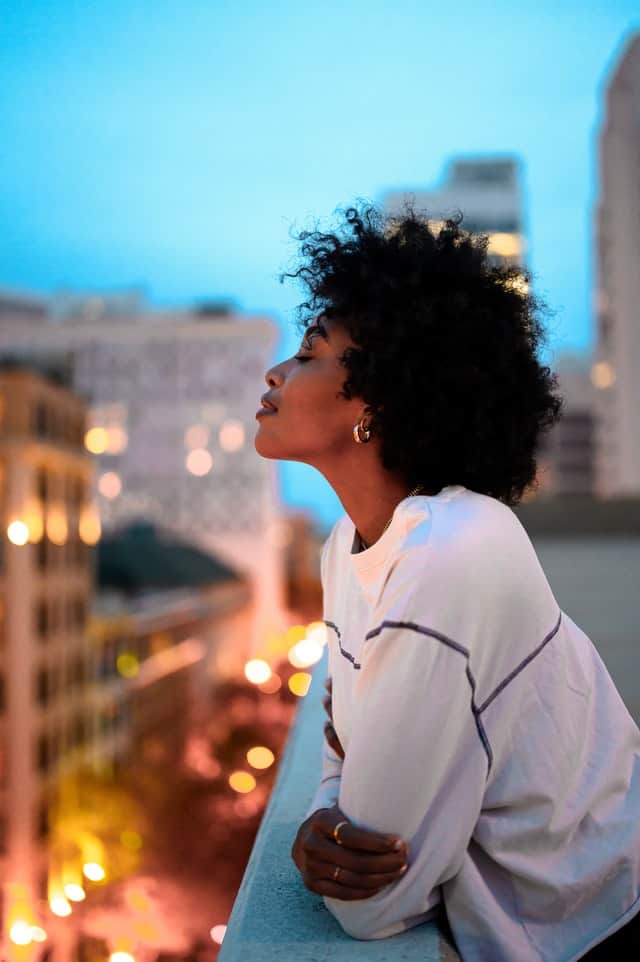 Woman with natural hair leaning over apartment balcony in the sunset, smiling.