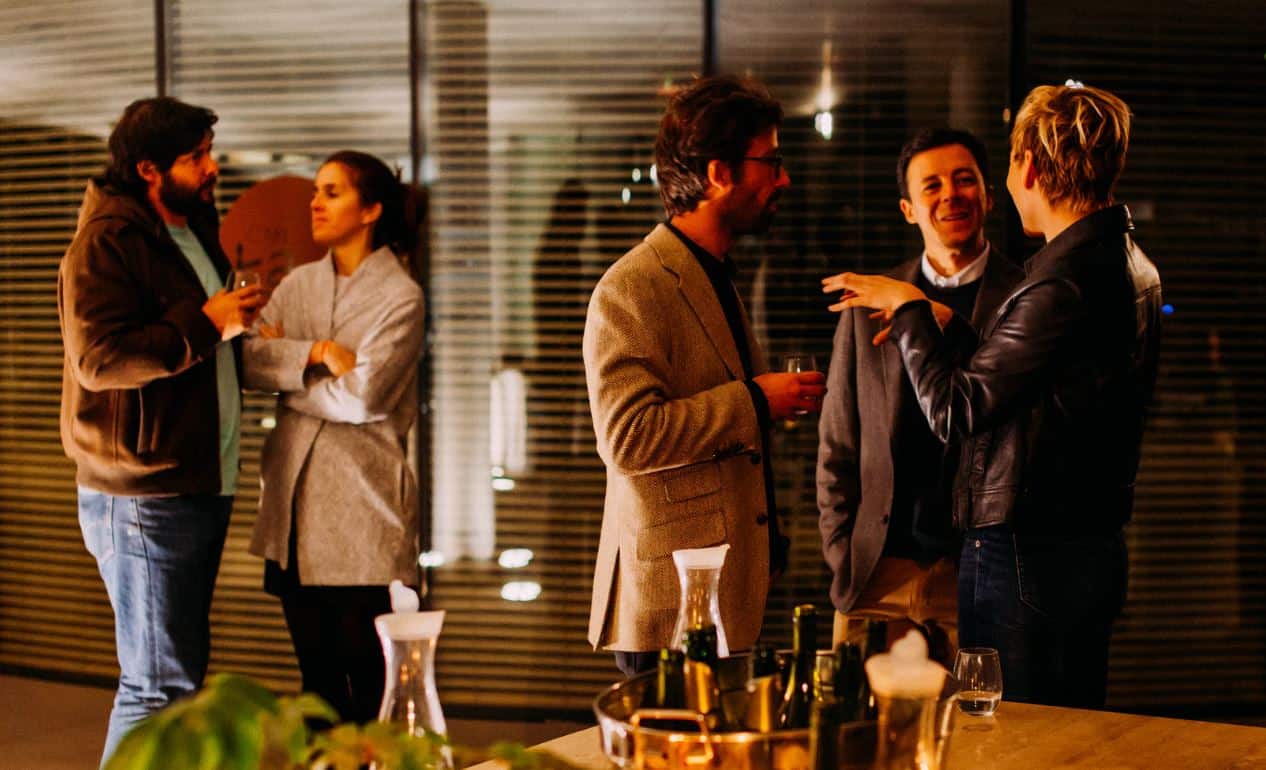 Five people in formalwear having a discussion in a setting that seems like an after-hours office get-together. There are drinks on the table at the forefront.