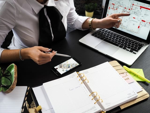 Woman in formalwear pointing at a property plan on both a laptop and phone screen, with other files and notes on the desk in front of her.