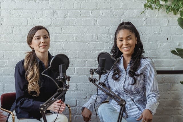 Two women sitting in front of microphones and smiling, preparing for a radio show or podcast.