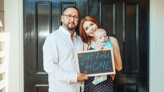 Man and woman holding a chalkboard and a baby, smiling and standing in front of a house door. The chalkboard has little harts drawn on it and &quot;our first home&quot; written out.