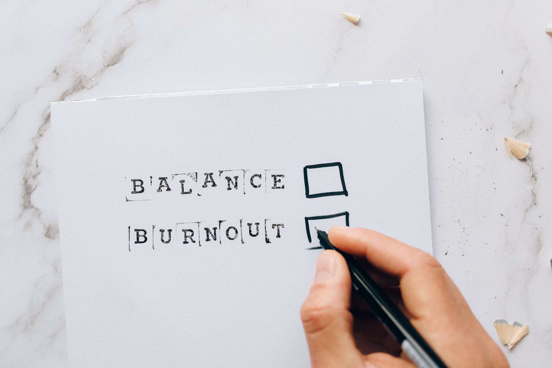Picture representing the importance of time management: A piece of paper on a marbletop table saying "Balance" and "Burnout" with a tick box next to each. A hand with a pen is hovering over the tickbox next to "Burnout". On the right side of the image, there are pencil shreds all over the table.