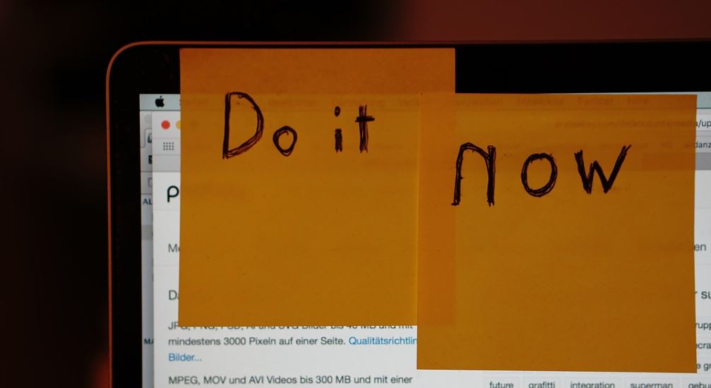 Two yellow post-it notes stuck to the upper right corner of a laptop screen. One of them has "do it" scribbled with a marker, while the second one more towards the middle of the screen says "now" in bigger lettering.