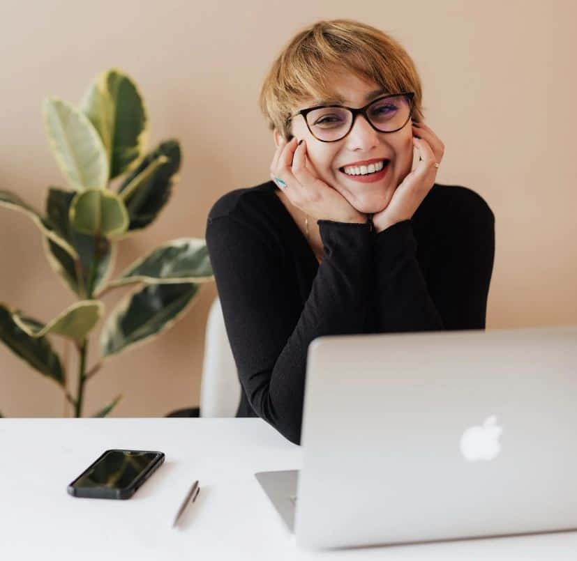 Woman sitting at desk, resting elbows on the table and her face in her palms, smiling. On the desk, there's a laptop, a phone, a pen, and a ficus plant in the background.
