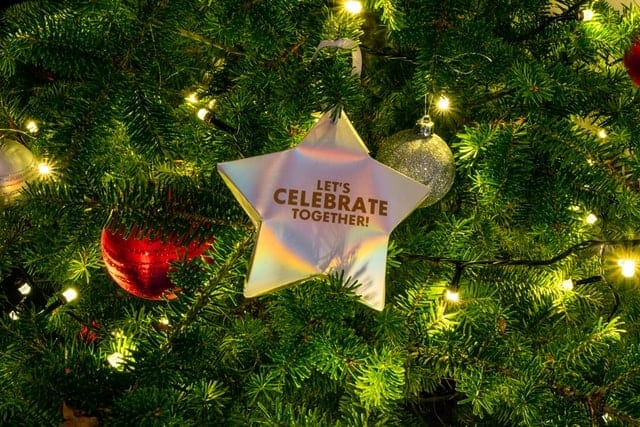 Star ornament in branches of Christmas tree with dim yellow lights, with text on it saying "Let's celebrate together".